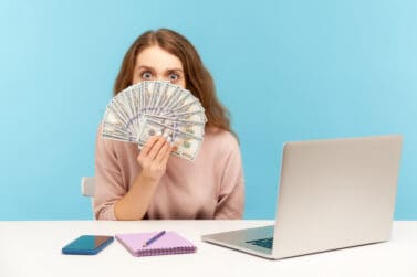 how to make quick money in one day Funny surprised woman employee sitting at workplace with laptop and peeking out of dollar banknotes, looking with amazed big eye, shocked by high salary. indoor studio shot isolated, blue background